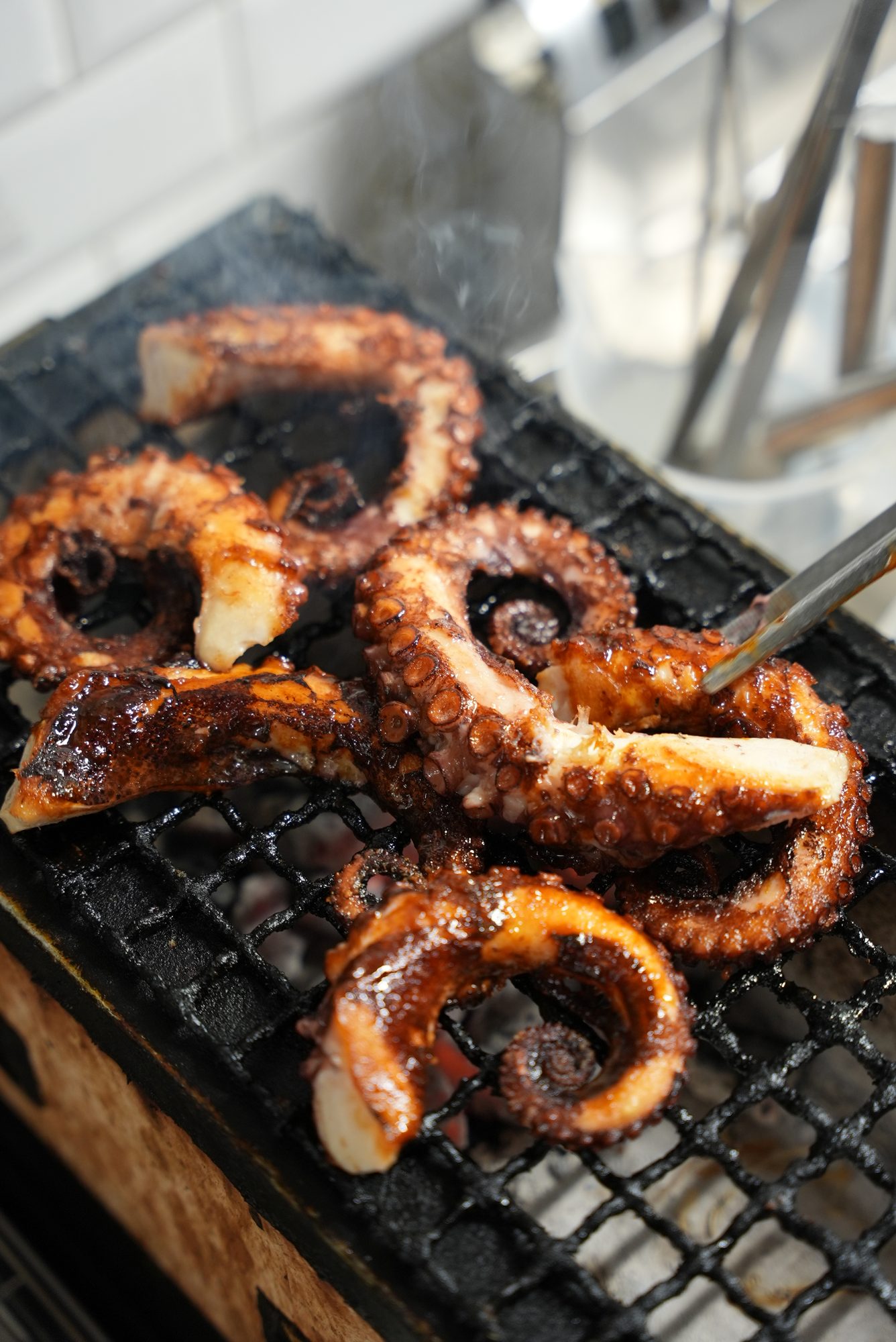 Tentacoli di polpo glassati sulla griglia, sopra la brace accesa, con il fumo che sale e una pinza.