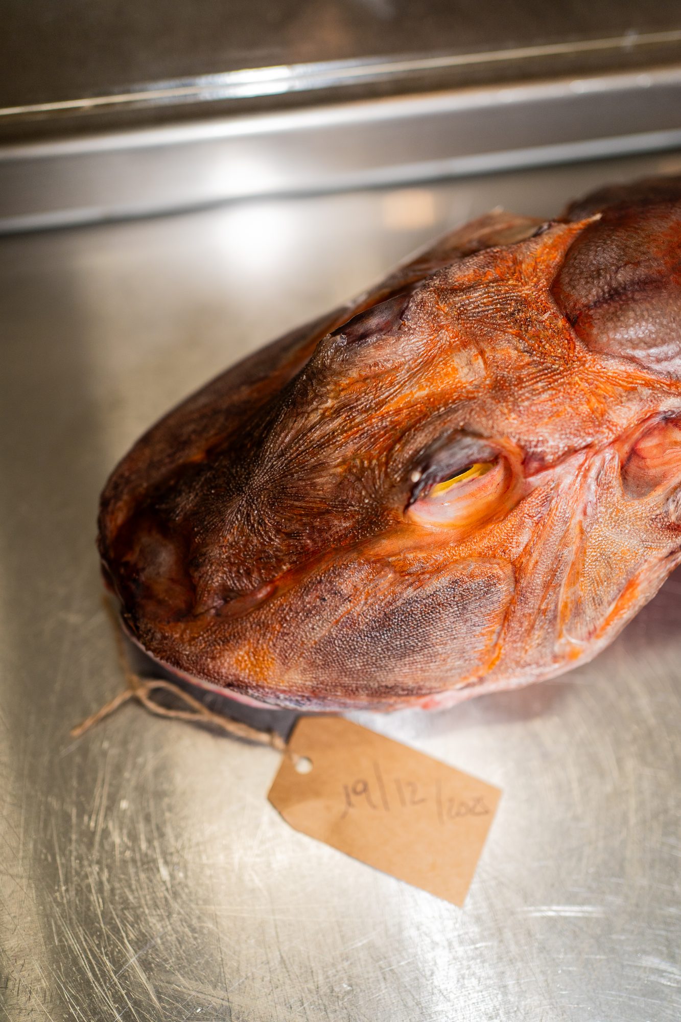 La testa di un pesce con un cartellino scritto a mano, legato con lo spago, su un vassoio d’acciaio.